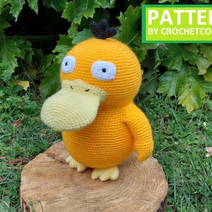 Puede incluir: Un peluche de Psyduck hecho a mano, tejido a ganchillo en amarillo brillante con un pico y pies amarillo claro. El peluche tiene ojos blancos y azules y un pequeño mechón de pelo negro. El texto "PATTERN BY CROCHETCONI" está incluido.