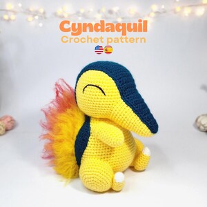 May include: A handmade Cyndaquil crochet pattern. The plush toy is yellow with a navy blue snout and back markings. It has a fluffy, orange and yellow tail. The text "Cyndaquil Crochet pattern" is displayed above the toy.