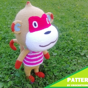 Pode incluir: Uma boneca de macaco de crochê com corpo bege, mãos e pés vermelhos e rosto branco. O macaco usa uma camisa listrada rosa e branca e uma máscara vermelha. A imagem inclui o texto "PATTERN BY CROCHETCONI" em um canto verde e amarelo.
