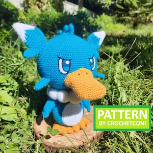 May include: A handmade blue, white, and orange crochet plush toy of a cartoon duck-like creature. The toy has large eyes, a yellow beak, and white accents. The text "PATTERN BY CROCHETCONI" is in a green box.