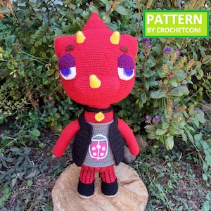 Könnte beinhalten: Handgemachte rote Häkelpuppe mit gelben Hörnern, lila Augen und einer schwarzen Weste. Die Puppe trägt ein graues Hemd mit Marienkäfer-Design und eine rot karierte Hose. Der Text "PATTERN BY CROCHETCONI" steht in einem grünen Rechteck.