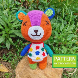 May include: A handmade orange crochet bear doll with colorful accents. The bear has a white face, black stitched eyes, and a stitched mouth. It wears a white top with colorful star shapes. The text "PATTERN BY CROCHETCONI" is in a green box.