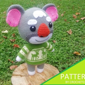 May include: A crocheted koala doll with a gray head, pink ears, and a red nose. It wears a green and white sweater with a deer pattern. The doll is standing on a wooden stump in a grassy area. The image includes the text "PATTERN BY CROCHETCONI".
