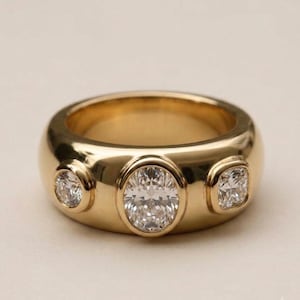 Könnte beinhalten: Ein Ring aus poliertem Gelbgold mit drei eingefassten, klaren Edelsteinen. Der mittlere Stein ist ein großer Diamant im Ovalschliff, flankiert von zwei kleineren Diamanten im Rundschliff, die bündig in die glatte, abgerundete Oberfläche eingelassen sind.
