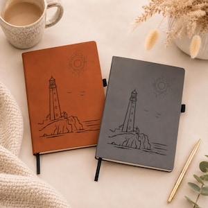 May include: Two rectangular notebooks, one in tan and one in dark gray, each featuring a black line art illustration of a lighthouse on a rocky cliff with a sun and birds. Both notebooks have a black elastic band closure attached to the side.