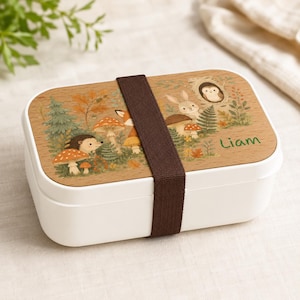 May include: A rectangular white bento box with a wood-grain lid featuring a forest illustration of a hedgehog, rabbit, owl, and mushrooms. A dark brown elastic band secures the lid, and the name Liam is printed in green on the right side.