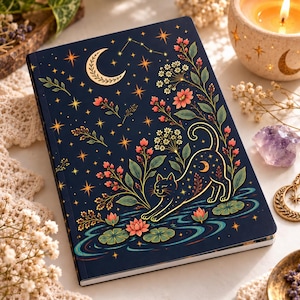 May include: A dark blue notebook featuring a gold-toned illustration of a cat stretching near a pond with lotus flowers. The cover design includes a crescent moon, stars, and floral sprigs in shades of pink, green, and gold, creating a detailed scene.