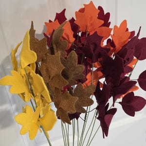 May include: A collection of artificial autumn leaves crafted from felt in shades of yellow, brown, orange, and deep burgundy. Each leaf is attached to a thin, flexible green wire stem, creating a bouquet of foliage with varied shapes.