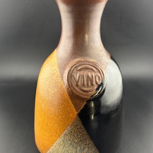 May include: A ceramic wine bottle with a brown neck and a body divided into three sections: orange, grey, and black. The word "VINO" is embossed on the front. The bottle has a narrow neck and a rounded base.