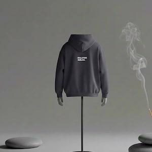 Può includere: Una felpa con cappuccio grigio scuro con la scritta bianca "ISOLATION BUILDS." sul retro. La felpa con cappuccio è esposta su un supporto per manichino nero su sfondo grigio. Il fumo dell'incenso si alza sullo sfondo.