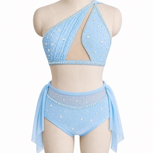 May include: Pale blue dance costume with rhinestone detailing. The two-piece set features a one-shoulder top and high-waisted shorts. A matching choker and sheer fabric accents complete the look. The costume is displayed on a mannequin.