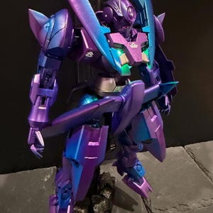 May include: A robotic action figure featuring a metallic purple and teal color-shifting finish. The figure has a pointed head design, articulated limbs, and a chest plate with a glowing green light. It stands on a dark, textured base.