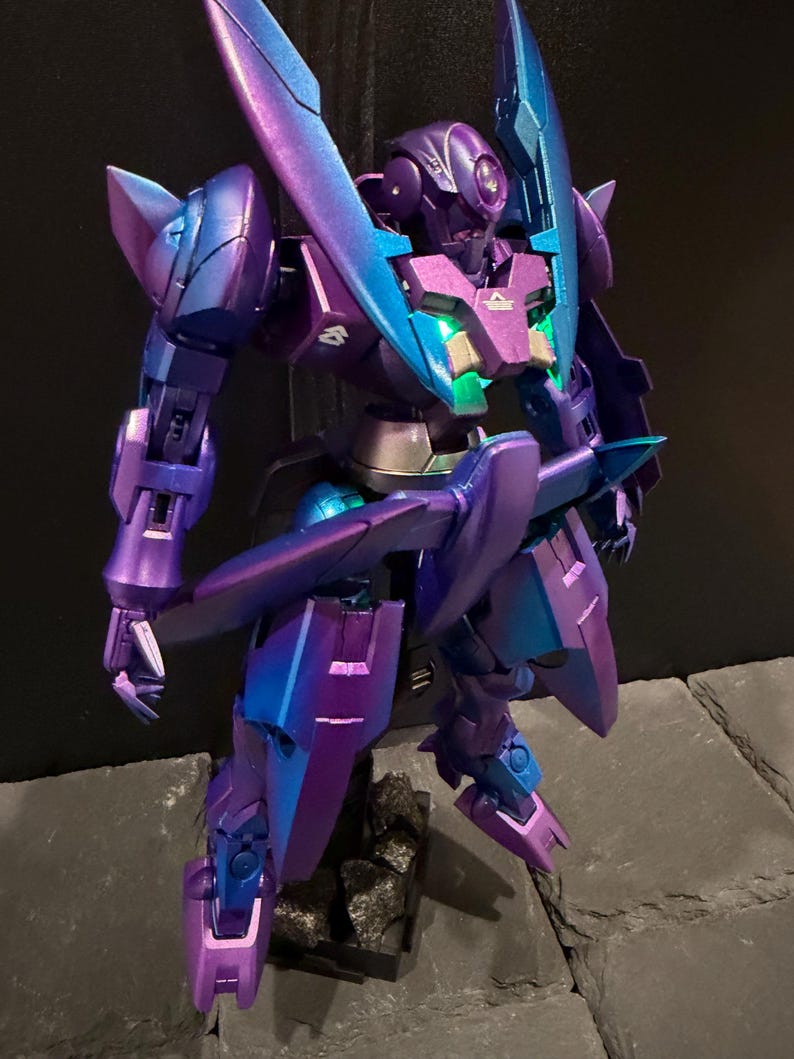 May include: A robotic action figure featuring a metallic purple and teal color-shifting finish. The figure has a pointed head design, articulated limbs, and a chest plate with a glowing green light. It stands on a dark, textured base.
