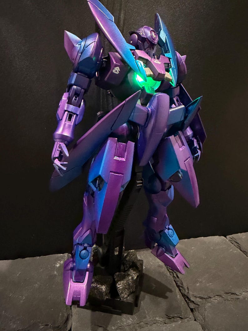 May include: A posable robot action figure featuring a metallic purple and blue color-shifting finish. The figure has sharp, angular armor plating, pointed shoulder guards, and a glowing green light integrated into the center of its chest piece.
