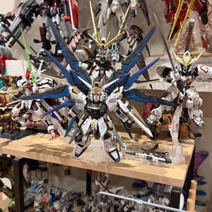 May include: A collection of detailed, multi-colored Gundam model kits displayed on a wooden shelf. The central model features white, blue, and gold accents with large, stylized wings. Other models in the background showcase various designs and color schemes.