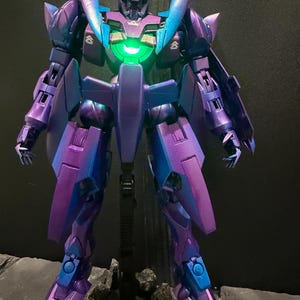 May include: A standing robot action figure featuring a metallic purple and blue color-shifting finish. The figure has a glowing green circular light in the center of its chest, angular armor plating, and small white insignia markings on the shoulders.