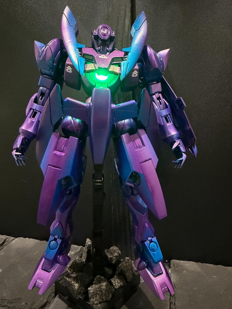 May include: A standing robot action figure featuring a metallic purple and blue color-shifting finish. The figure has a glowing green circular light in the center of its chest, angular armor plating, and small white insignia markings on the shoulders.