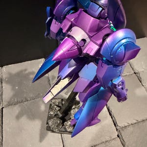 May include: A robotic action figure featuring a metallic iridescent finish that shifts between deep purple and vibrant blue. The model has a complex, angular design with pointed shoulder armor, a central cone-shaped chest piece, and articulated limbs.