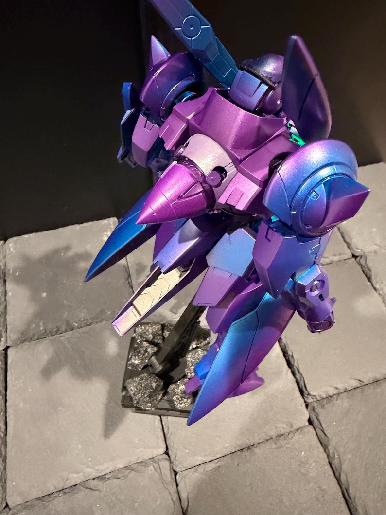 May include: A robotic action figure featuring a metallic iridescent finish that shifts between deep purple and vibrant blue. The model has a complex, angular design with pointed shoulder armor, a central cone-shaped chest piece, and articulated limbs.