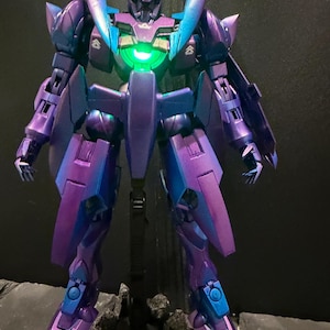 May include: A robotic action figure with a metallic purple and blue gradient finish. The figure features a glowing green circular light in the center of its chest, articulated limbs, and pointed shoulder armor, standing on a dark base.