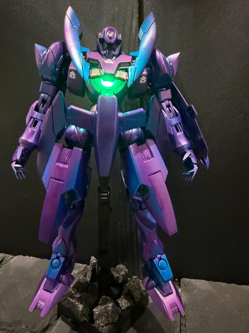 May include: A robotic action figure with a metallic purple and blue gradient finish. The figure features a glowing green circular light in the center of its chest, articulated limbs, and pointed shoulder armor, standing on a dark base.