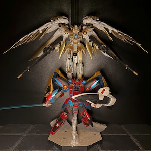 May include: Two detailed robot action figures. The top figure is primarily white and gold with large, articulated wings. The bottom figure is red, blue, and black, holding a sword and shield. Both figures are posed against a dark background.