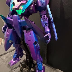 May include: A robotic action figure featuring a metallic purple and blue color scheme with a glowing green chest piece. The figure has articulated limbs, sharp angular armor plating, and a small white emblem on the shoulder, standing on a base.