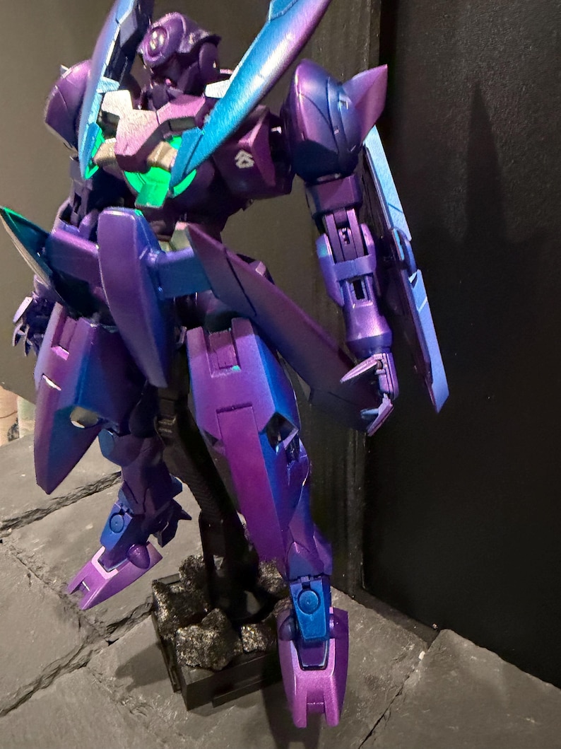 May include: A robotic action figure featuring a metallic purple and blue color scheme with a glowing green chest piece. The figure has articulated limbs, sharp angular armor plating, and a small white emblem on the shoulder, standing on a base.