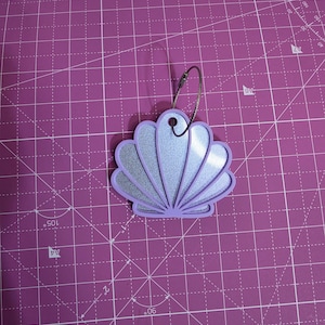 May include: A lavender seashell-shaped ornament featuring a shimmering silver glitter inlay. The shell has a cutout design with a circular hole at the top, through which a thin, flexible metal wire loop is threaded for hanging purposes.