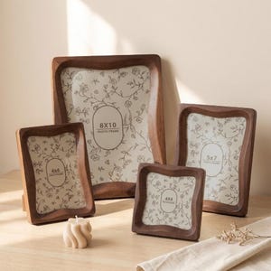 Könnte beinhalten: Eine Sammlung von fünf Holz-Fotorahmen in verschiedenen Größen. Die Rahmen haben eine dunkelbraune Oberfläche und ein wellenförmiges Kantendesign. Die Rahmen enthalten floral gemusterte Einsätze mit den Größen 4x4, 4x6, 5x7 und 8x10.