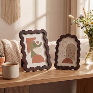 Puede incluir: Dos marcos de fotos decorativos de madera con borde ondulado. Los marcos contienen impresiones artísticas abstractas en tonos naranja, verde y beige. Una impresión presenta un diseño de sol y luna. Los marcos se exhiben sobre una superficie de madera.
