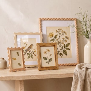 Peut inclure: Trois cadres photo en bois de tailles variées avec des bords festonnés et torsadés. Les cadres contiennent des illustrations botaniques et d'oiseaux sur papier crème. Ils sont disposés sur une surface plane avec un vase en céramique.