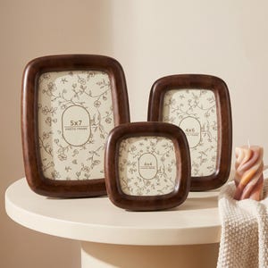 May include: Three wooden photo frames in varying sizes, with rounded corners and a dark brown finish. The frames hold floral patterned inserts and are labeled 5x7, 4x6, and 4x4. A decorative candle sits nearby.