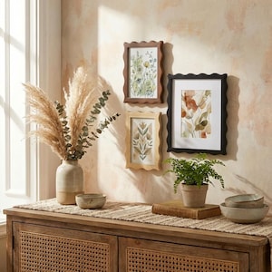 May include: Three framed botanical art prints with scalloped edges hang on a wall. The frames are brown, black, and light wood. A vase with dried pampas grass and a small potted plant sit on a wooden cabinet.