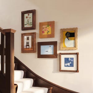 May include: A collection of framed art pieces in various styles and sizes, mounted on a wall. The frames are made of wood and feature different designs, including beaded and scalloped edges. The artwork includes floral, abstract, and portrait images.