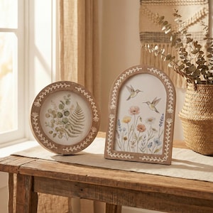 May include: Two wooden framed artworks. One is round with a botanical print of green leaves. The other is arched, featuring hummingbirds and colourful flowers. Both frames have a white leaf design around the edges. They sit on a wooden table.