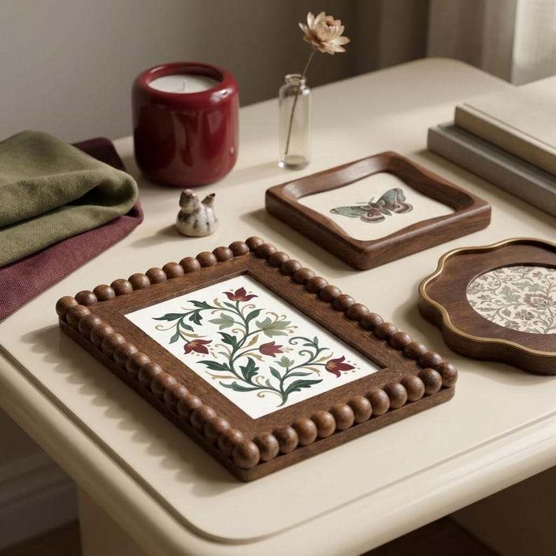 May include: A collection of decorative items on a cream-colored surface. Includes a dark red candle holder, framed botanical art, a small ceramic bird, and a tray with a butterfly design. The frames have wooden beaded edges.