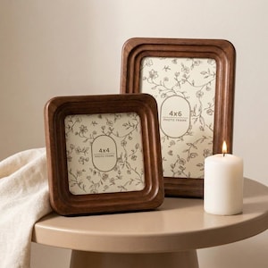 May include: Two wooden photo frames, one square and one rectangular, with floral patterned inserts. The frames are a rich brown colour and have rounded edges. A white candle with a lit flame sits nearby. The square frame is labelled "4x4 PHOTO FRAME", and the rectangular frame is labelled "4x6 PHOTO FRAME".