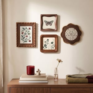 May include: A collection of framed art pieces with botanical and butterfly illustrations. The frames are made of dark wood and vary in shape. A red candle, small vase, and stacked books sit on a wooden surface below the art.