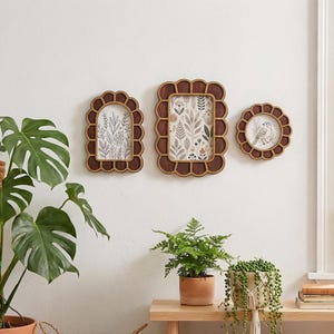 Peut inclure: Trois cadres photo décoratifs en bois aux bords festonnés sont montés sur un mur blanc. Les cadres sont de formes variées : en arc, rectangulaire et circulaire. Chaque cadre contient des impressions d'art botanique aux tons neutres. Des plantes vertes sont au premier plan.