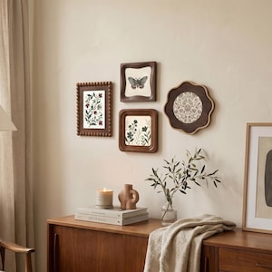 May include: A collection of framed botanical art and a decorative wall display. The frames are various shapes and sizes, with brown wooden frames. The artwork includes floral and butterfly prints. A candle, vase, and books are on a wooden cabinet below the art.