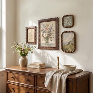 May include: A wooden dresser holds a beige ceramic vase with wildflowers, a stack of books, a brass candlestick, and a bowl. Three dark brown beaded frames hang on the wall, displaying a floral bouquet, a botanical sketch, and a coastal landscape.