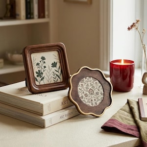 May include: Two decorative wooden picture frames, one square and one scalloped, with floral artwork. The square frame displays a botanical print, while the scalloped frame features a patterned design. A lit red candle and a stack of books are also visible.