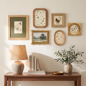 Könnte beinhalten: Eine Galeriewand mit sechs gerahmten botanischen und Landschaftsbildern, eine Tischlampe mit strukturiertem Fuß und Leinenschirm, ein Bücherstapel, eine kleine goldene Schale und ein Olivenbaum im Topf auf einem Konsolentisch aus Holz.
