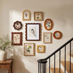 Puede incluir: Una pared de galería con diez marcos de fotos variados en diferentes formas, incluyendo diseños rectangulares, ovalados, arqueados y festoneados, que muestran ilustraciones botánicas, de aves y de manos sobre una pared clara.