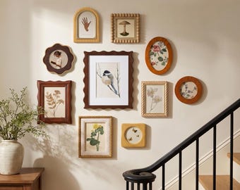 May include: A gallery wall arrangement featuring ten assorted picture frames in various shapes, including rectangular, oval, arched, and scalloped designs, displaying botanical, bird, and hand illustrations on a light-colored wall above a staircase.
