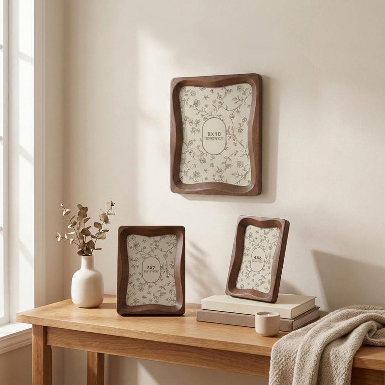 May include: Three dark brown wooden photo frames with wavy, organic edges. One frame is mounted on the wall, while two smaller frames stand on a surface. Each frame contains a cream-colored insert with a delicate floral line drawing pattern.
