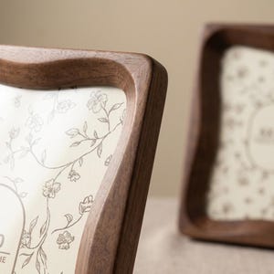 May include: Two rectangular picture frames crafted from dark brown wood with a smooth, curved edge profile. Each frame holds a cream-colored insert featuring a delicate, thin-lined botanical illustration of flowers and vines in a dark brown ink.