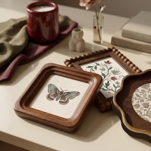 May include: Three wooden picture frames with different designs. One frame features a butterfly illustration, another has a floral print, and the third has a patterned design. A red candle, a small vase with a flower, and a small sculpture are also visible.