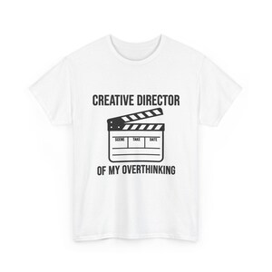 May include: A white short-sleeved t-shirt featuring a black graphic of a film clapperboard in the center. Above the clapperboard, the text reads CREATIVE DIRECTOR, and below the clapperboard, the text reads OF MY OVERTHINKING in a bold font.